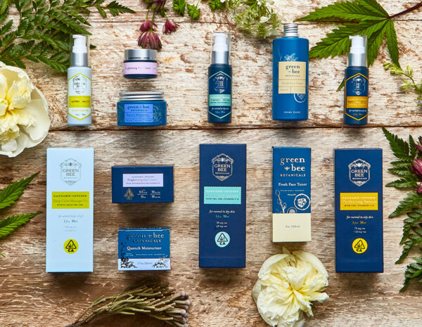 Photo of six Green Bee Botanicals cannabis-infused skincare and hemp-infused skincare products, including (left to right, top to bottom) Deep Calm Massage Oil, Brightening Eye Cream, Quench Moisturizer, Renewing Serum, Fresh Toner and Perfecting Serum. Product bottles and boxes are laying on a farm table surrounded by fresh flowers. Photo by Sonoma Hills Farm.
