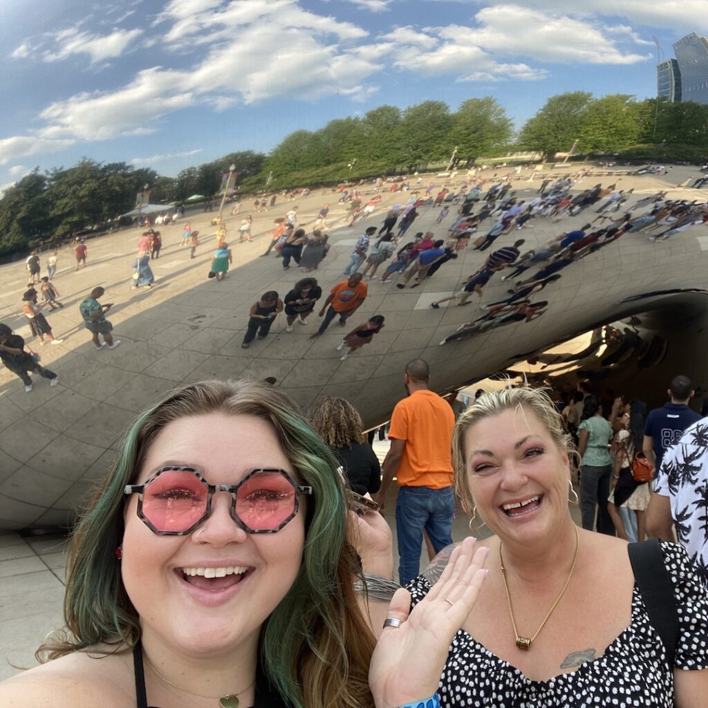 Photo of Jen Bean and her mom Kristi Bean laughing in front of the famous Chicago sculpture, Cloud Gate, known as The Bean. Reflected in The Bean are people, the park and the blue sky with white clouds