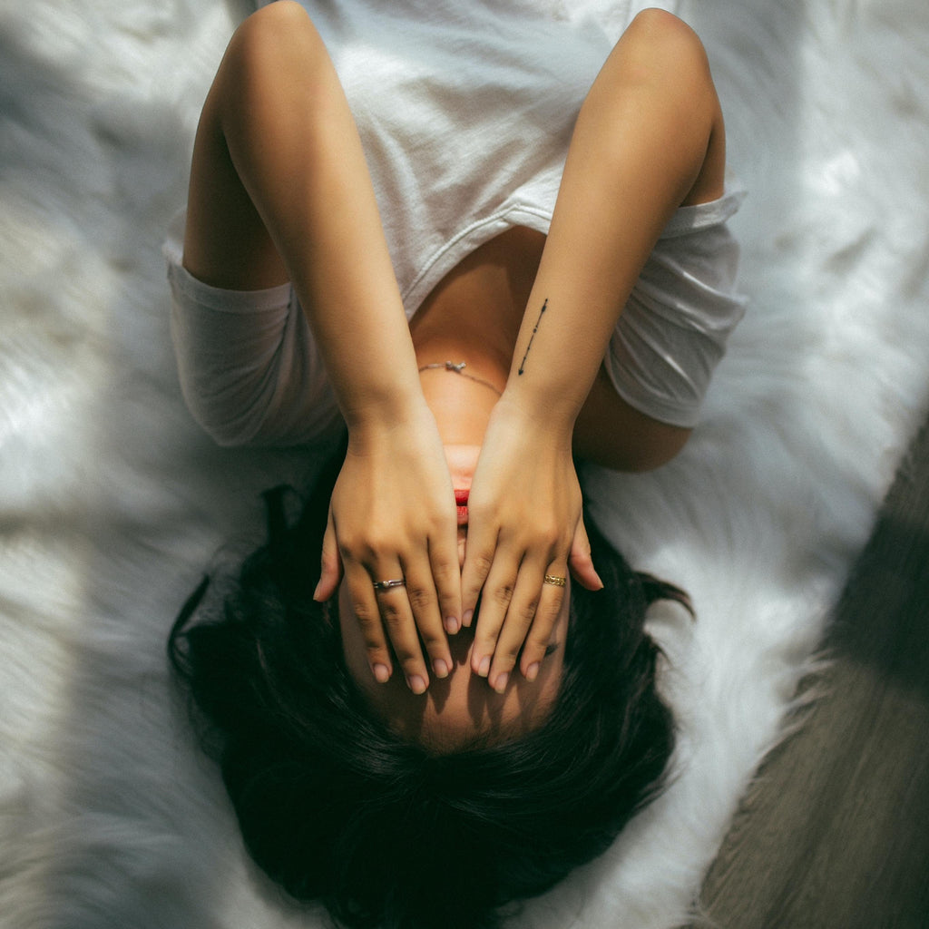 Photo from above of a young woman laying on a bed with her hands covering her face - dark hair - olive skin tone - small tattoo - rings -white tshirt -white sheets
